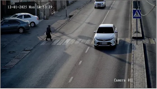 PED camera color view of a sedan stopped in a marked crosswalk beside a pedestrian sign, with a pedestrian walking nearby, timestamped and labeled Camera 01