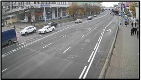 Overview (OV) camera wide shot of a city street with vehicles and pedestrians on the sidewalk, with a timestamp overlay