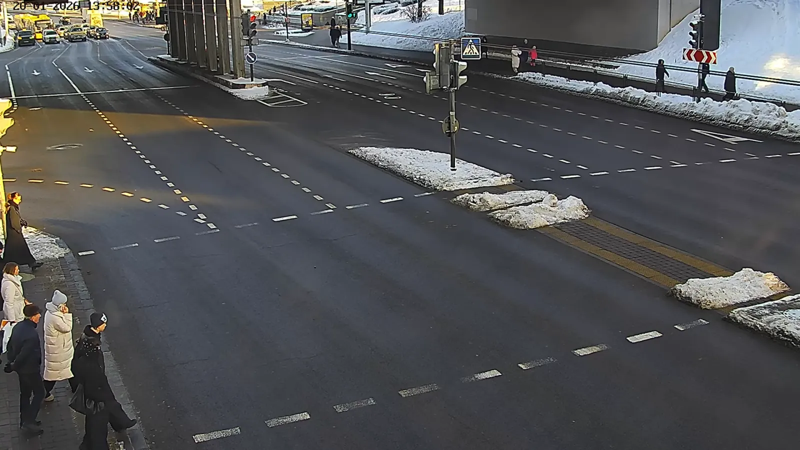 OV camera wide shot of a wide intersection beneath an overpass with pedestrians on the sidewalk and snow piled along the curb