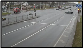 TL camera view from beneath an overpass showing a vehicle at the stop line, an illuminated traffic light, and pedestrians on the sidewalk in the rain