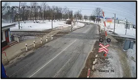 OV camera wide shot of a road crossing railway tracks with a crossbuck signal, barrier posts, and open snow-covered fields on both sides